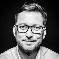 Black and white portrait of a smiling individual with short hair, wearing round glasses, and a casual t-shirt. The person has a friendly demeanor and is looking directly at the camera.