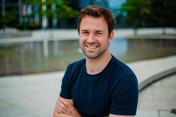 A person with short brown hair and a beard smiling at the camera, wearing a dark blue t-shirt and crossing his arms. The background is blurred with hints of greenery and an urban setting.