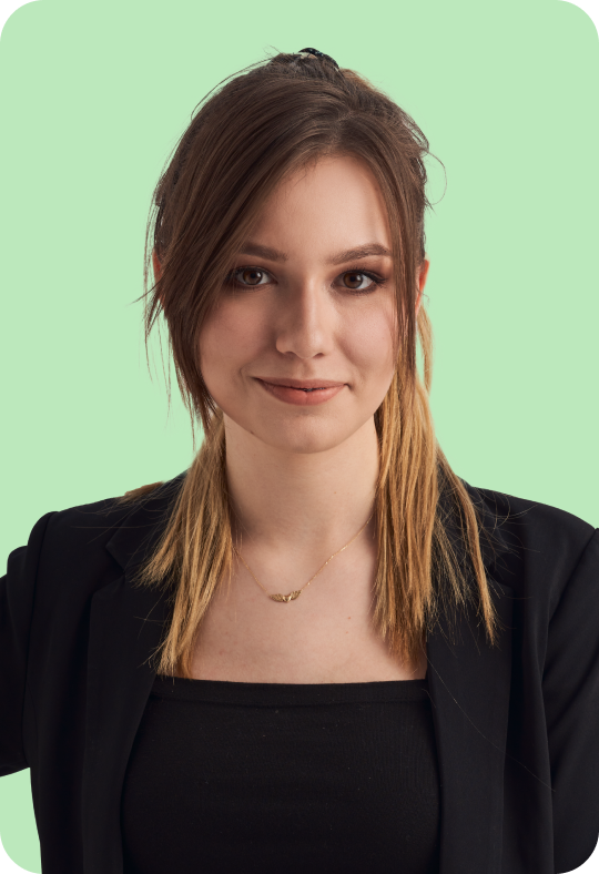 A person with shoulder-length straight brown hair, wearing a black blazer over a black top, giving a confident smile, against a pale green background.