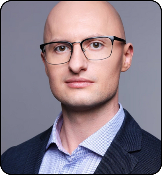 Portrait of a person with a bald head, wearing black-rimmed glasses, a blue checkered shirt, and a dark blazer. The individual has a neutral expression and is looking directly at the camera, set against a blurred grey background.