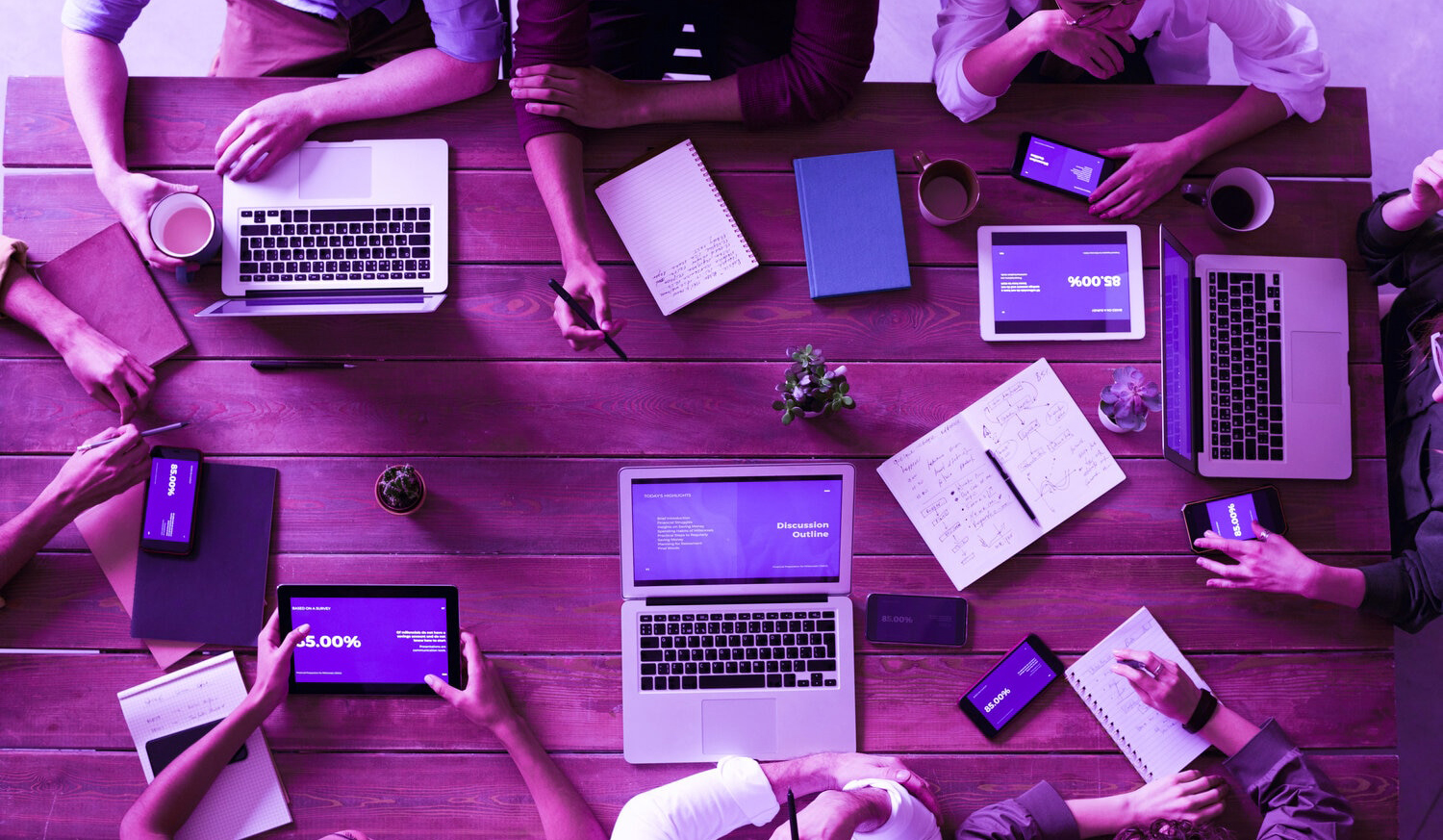 A photo depicting a table with multiple electronic devices, laptops, tablets, phones. There are people sitting at the table but they're faces are not visible. Visualises groupwork.